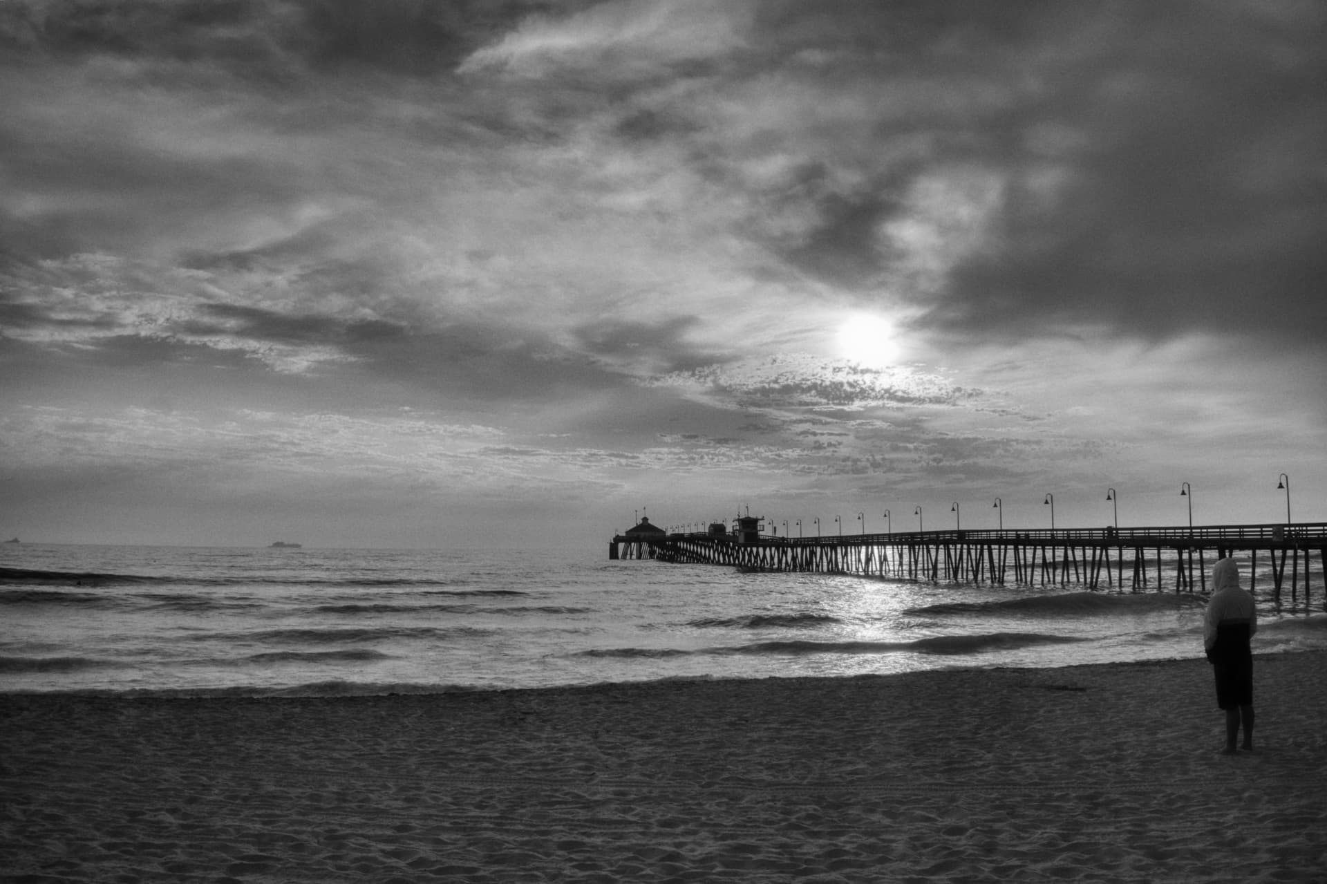 A hooded figure on a wide shore beneath an enormous sky, a distant pier behind